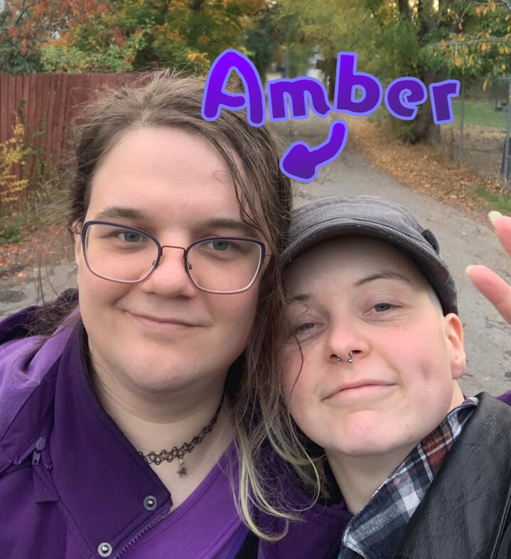 The left half of this chaos gremlin duo ;) selfie of Amber and her partner in front of an autumn alleyway with trees. Amber is wearing catseye glasses and is wrapping her arm around her partner doing a peace sign.